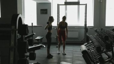 Göz kamaştırıcı fitness kızları yan yana duruyor ve ağır halterleri kaldırarak antrenman yapıyorlar. Spor salonunda vücut şekline sahip genç kadınlar birlikte spor yapıyorlar..