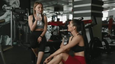 Spor kıyafetleri giymiş, antrenmandan sonra dinlenirken şişeden su içen güçlü vücutlu mutlu genç bayanlar. Yorgun, siyah saçlı kadınlar spor salonunda egzersiz aralarında ferahlatıyorlar..
