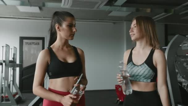 两名身材纤细 身穿运动服的女子在训练 喝水和使用智能手机后走在体育馆里 令人赞叹 锻炼和友谊的概念 — 图库视频影像