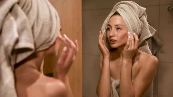 シャワー後のマッサージの動きと顔に保湿クリームを適用する自然の美しさを持つ美しい女性 バスルームで頭と体にタオルを身に着けている女の子 — ストック動画