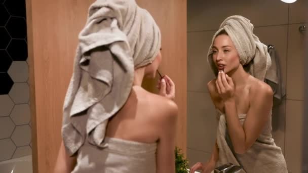 魅力的な女性は鏡を見てタオルに包まれ 朝のシャワー後口紅を適用します 幸せな女性は毎日のメイクや朝の儀式でバスルーム — ストック動画
