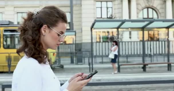 Вид сбоку девушки, использующей смартфон во время прогулки по улице — стоковое видео
