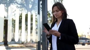 Şık takım elbiseli gülümseyen iş kadını sokakta yürüyor ve akıllı telefon kullanıyor. Güzel, koyu renk saçlı bir kadın, açık havada internette geziniyor..