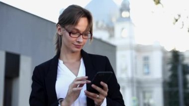 Gözlüklü, gülümseyen genç iş kadını dışarıdaki akıllı telefondan e-postalarını kontrol ediyor. Kahverengi saçlı güzel bir kadın internette resmi mektuplar yazıyor..