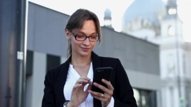 Resmi kıyafetli ve gözlüklü gülümseyen iş kadınının portresi şehir merkezinde dururken modern akıllı telefon üzerinde çalışıyor. Başarılı kariyer, teknoloji ve insanlar kavramı.