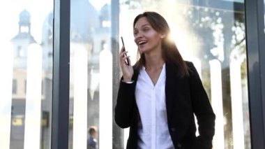 Takım elbiseli neşeli genç bayan sokakta dikiliyor ve akıllı telefondan konuşuyor. Kahverengi saçlı mutlu kadın açık havada konuşuyor..