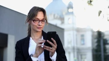 Açık havada duran ve akıllı telefondan iyi haberler okuyan resmi kıyafetli başarılı bir kadın. Uzaktan kumanda için modern cihaz kullanarak gözlüklü, gülümseyen esmer..