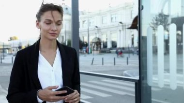 Resmi giyinmiş güzel bir kadın akıllı telefondan toplu taşıma zamanını kontrol ediyor. Kahverengi saçlı iş kadını otobüs durağında yürüyor ve modern aletler kullanıyor..