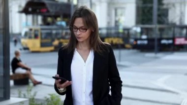 Gözlüklü ve ofis kıyafetli konsantre iş kadını büyük şehirde yürürken akıllı telefondan konuşuyor. Kahverengi saçlı çekici bir kadın cep telefonuyla konuşuyor..