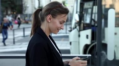 Otobüs durağında dikilirken, siyah ceketli, beyaz tişörtlü, gülümseyen bir kadının toplu taşıma programı araması. Açık havada modern akıllı telefon kullanan siyah saçlı çekici bir kadın..