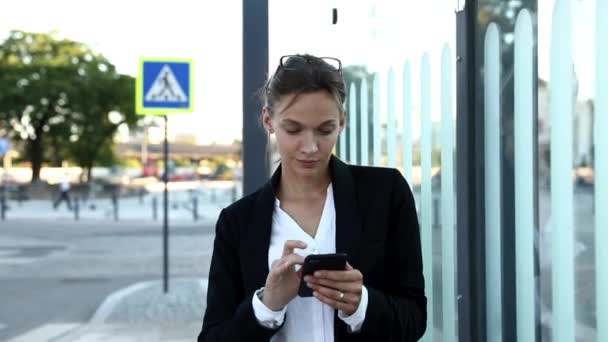 Tramvay istasyonunda oturan ve akıllı telefondan iş arkadaşlarıyla sohbet eden başarılı bir iş kadını. Açık havada çalışmak için interneti kullanan güzel bir kadın.. — Stok video