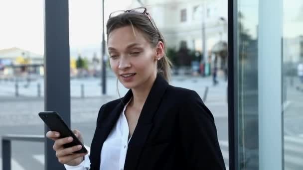 Şehir merkezinde dururken akıllı telefondan taksi çağıran güzel genç bir kadının portresi. Düğüme bağlanmış, gülümseyen ve kameraya bakan siyah saçlı mutlu kız.. — Stok video