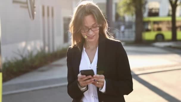 Pen dame i briller og formelle klær venter på en taxi mens hun står på gata. Forretningskvinne med brunt hår på mobil anvendelse utendørs. – stockvideo