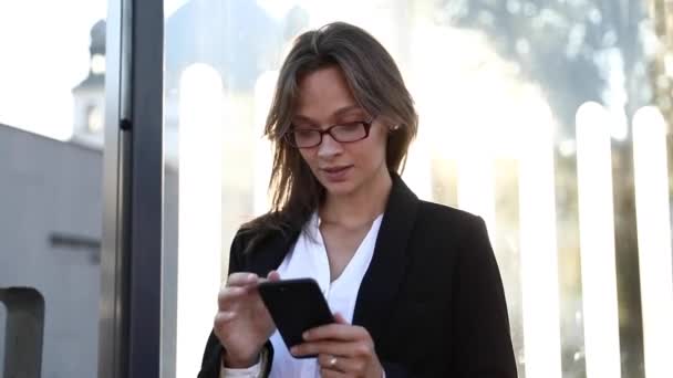 Gözlüklü ve ofis kıyafetli mutlu bir iş kadını, ulaşımı beklerken sosyal ağlarda arkadaşlarıyla sohbet ediyor. Çevrimiçi konuşma için akıllı telefonu kullanan güzel kadın. — Stok video