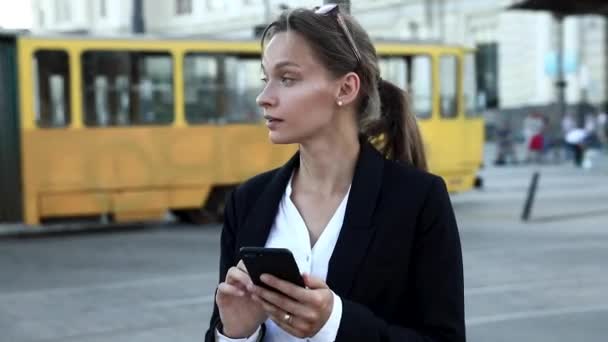 Mujer de negocios segura con camisa blanca y chaqueta negra de pie en la parada del tranvía y haciendo trabajo en línea en el teléfono inteligente. Joven dama con el pelo castaño atado en nudo usando gadget moderno al aire libre. — Vídeo de stock
