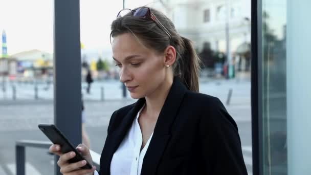Självsäker affärskvinna i formell klädsel lösa arbetsfrågor på smartphone när du står utomhus. Charmig dam med glasögon på mörkt hår står på stopp för kollektivtrafik. — Stockvideo