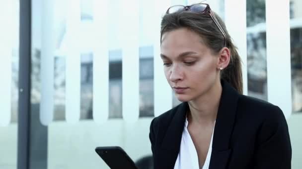 Femme d'affaires sérieuse en tenue formelle assis sur l'arrêt de bus et travaillant sur smartphone. Charmante dame aux cheveux foncés noués à l'aide d'un appareil moderne pendant le temps libre. — Video