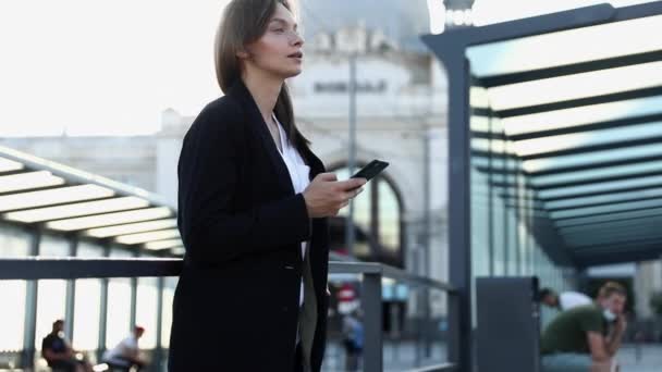 Mujer confiada en la ropa formal esperando el transporte público en la estación y trabajando en línea en el teléfono inteligente. Concepto de personas y tecnología moderna. — Vídeo de stock