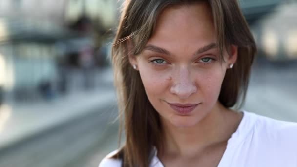 Närbild av glad ung kvinna med brunt hår poserar på kameran på järnvägsstationen. Vacker dam med blå ögon som står utomhus och ler. — Stockvideo