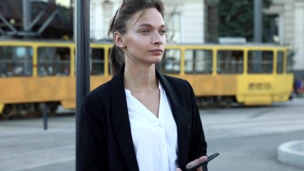 Atrakcyjna kobieta z brązowymi włosami związana węzłem stojąca na przystanku tramwajowym i czekająca na transport publiczny. Młoda dama w ubraniu casual gospodarstwa nowoczesny smartfon i patrząc na bok. — Wideo stockowe