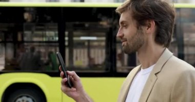 Resmi olarak gülümseyen sakallı adamın yan görüntüsü otobüs beklerken akıllı telefondan mesaj yazması. İnsan kavramı ve modern aygıt.