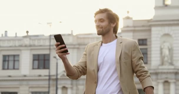 Příjemný mladý muž v obleku pomocí GPS navigace na moderním smartphonu. Muž turista s kufříkem hledající směr v mobilní aplikaci. — Stock video