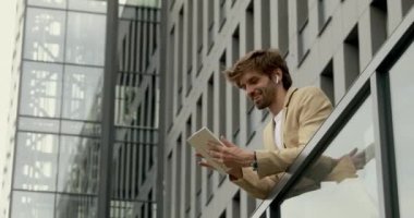 Ofisin balkonunda dikilirken dijital tablet kullanan takım elbiseli yakışıklı sakallı adam. Güven, kariyer ve teknoloji kavramı.