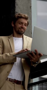 Resmi giyinmiş neşeli genç adam ofis merkezinin yanında duruyor ve modern dizüstü bilgisayarını iş için kullanıyor. İnsan kavramı, teknoloji ve güven.