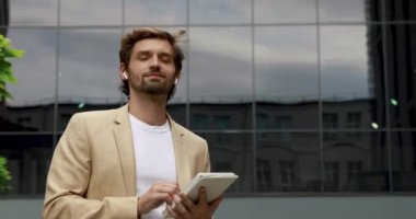 Resmi kıyafetli mutlu yakışıklı adam iş merkezinin yanında dikiliyor ve dijital tablet kullanıyor. Kablosuz kulaklıklı gülümseyen adam temiz havada duruyor ve modern cihaz üzerinde çalışıyor..