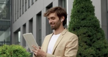 Şık giyinmiş sakallı bir adam iş merkezinin yanında dikilirken dijital tablet ekranına bakıyor. Yakışıklı adam modern aletlerle boş zamanların tadını çıkarıyor..