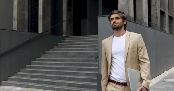 Boldog szakállas férfi vezeték nélküli laptoppal sétál az irodaház közelében. Jóképű üzletember hivatalos ruházatban időt tölt a szabadban modern eszközzel.