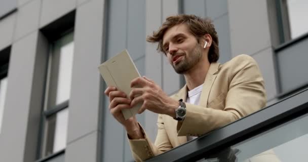 Low angle view of handsome man in business suit having video chat on digital tablet while standing outdoors. Concept of online communication and modern technology. — Stock Video