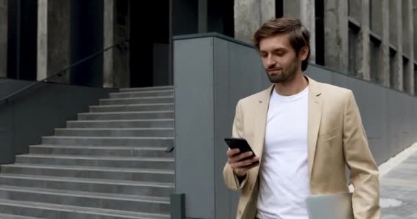 Уверенный молодой бизнесмен в официальной одежде гуляет по городу, пользуется современным смартфоном и держит в руках ноутбук. Концепция людей и современного оборудования. — стоковое видео