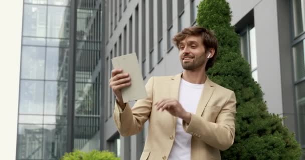 Hombre guapo en traje de negocios hablando y haciendo gestos durante una videollamada en una tableta digital. Varón barbudo en auriculares de pie al aire libre y tener conversación en línea. — Vídeos de Stock