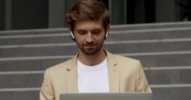 Homem barbudo feliz usando laptop sem fio, sentado nas escadas, sorrindo e olhando para a câmera. Bonito empresário em roupas formais e fones de ouvido modernos trabalhando fora do escritório. — Vídeo de Stock