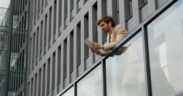 Atraktivní mladý muž ve formálním oblečení pomocí bezdrátových sluchátek a digitálního tabletu pro videohovory venku. Pohledný muž pracující na moderním zařízení. — Stock video