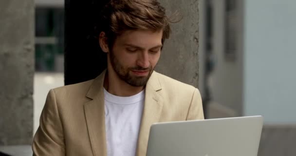 Портрет улыбающегося бизнесмена в стильном костюме, просматривающего интернет в портативном компьютере на открытом воздухе. Счастливый бородатый парень работает удаленно на современном ноутбуке. — стоковое видео