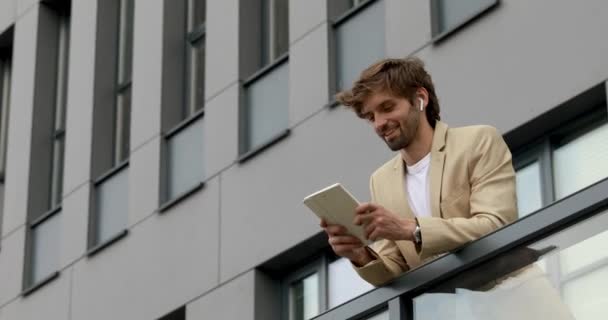 Joven hombre de negocios sonriente en auriculares inalámbricos usando tableta digital durante el tiempo libre. Hombre relajado en ropa formal de pie fuera con un dispositivo moderno. — Vídeo de stock