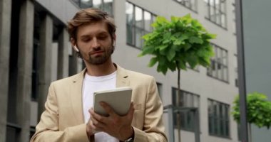 Kablosuz kulaklıklı, gülen sakallı adam dijital tabletli, modern gri binanın yanında duruyor. Başarılı kariyer ve aletler kavramı.