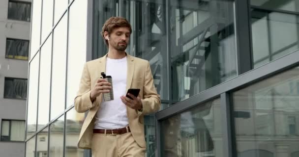 Ευτυχισμένος όμορφος άντρας με επίσημο ένδυμα πίνει καφέ από το θερμικό κύπελλο και χρησιμοποιεί smartphone κατά τη διάρκεια του ελεύθερου χρόνου. Γενειοφόρος τύπος φορώντας ασύρματα ακουστικά ενώ χαλαρώνει από την εργασία σε εξωτερικούς χώρους. — Αρχείο Βίντεο