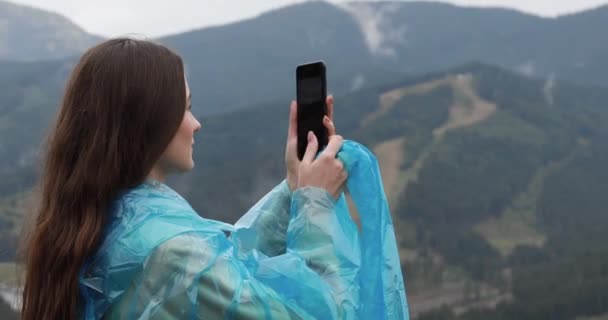 Nainen turisti sininen sadetakki ottaa valokuvan älypuhelimella vuorten joukossa pilviä. Houkutteleva tyttö käyttää modernia laitetta vaelluksen aikana luonnossa. — kuvapankkivideo