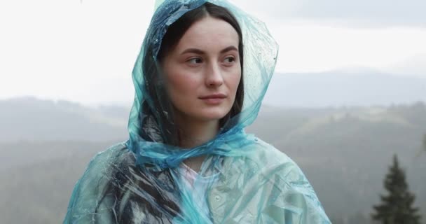 Retrato de una hermosa niña en impermeable azul con capucha entre las montañas durante el tiempo lluvioso. Mujer joven con el pelo castaño sonriendo y mirando a la cámara. — Vídeos de Stock