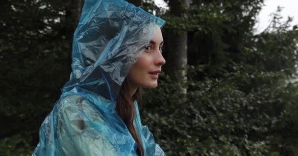 파란 비옷을 입은 매력적 인 젊은 여성이 비오는 날씨에 산 정상에 후드를 쓴 채서 있습니다. 과장 된 개념, 관광 및 평온 함의 개념. — 비디오