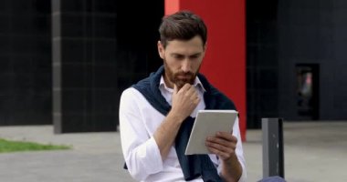 Ciddi yüz ifadesi olan sakallı bir adam sokakta oturup dijital tablet kullanıyor. Genç erkek, modern aletlerin yardımıyla uzaktan çalışıyor..