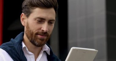 Dijital tablet ekranına bakıp gülümseyen şık saç kesimli ve sakallı genç adamı yakından çek. Beyaz gömlekli yakışıklı adam, temiz havanın tadını çıkarıyor..