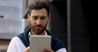 Beyaz tişörtlü gülümseyen sakallı adam iş merkezinin yanında otururken dijital tablet üzerinde çalışıyor. Modern teknolojiyi ve uzaktan çalışmayı kavra.