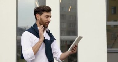 Beyaz gömlekli yakışıklı adam, modern binanın yanında dikilirken video görüşmesinde çalışıyor. Sakallı erkek temiz hava için dijital tablet kullanıyor..