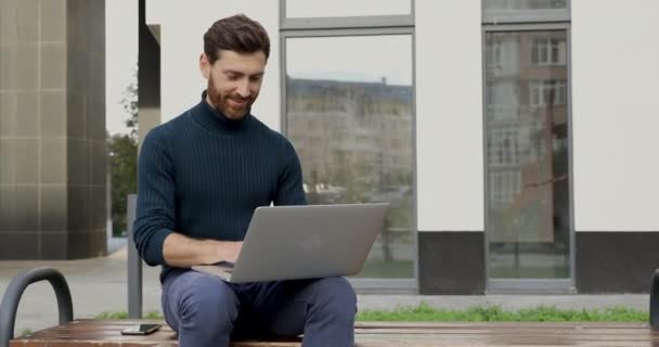 Sonriente hombre barbudo que trabaja en el ordenador portátil mientras está sentado en el banco cerca de un edificio moderno. Guapo masculino navegación por Internet en el ordenador portátil al aire libre. — Vídeos de Stock