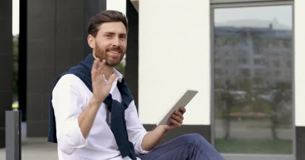 Hombre atractivo con barba saludando a alguien con la mano mientras está sentado al aire libre con la tableta digital. Concepto de personas, gestos y tecnología moderna. — Vídeos de Stock