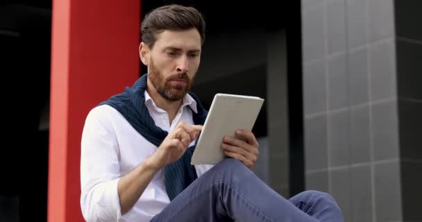 Gündelik giysiler içinde başarılı bir iş adamı dijital tablet üzerinde uzun çalışmaktan yorulmuş hissediyor. Sakallı erkek ofis binasının yanında oturuyor ve esniyor.. — Stok video